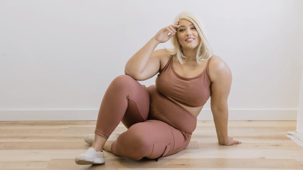 Person sitting on a wooden floor in a workout outfit smiling at the camera.