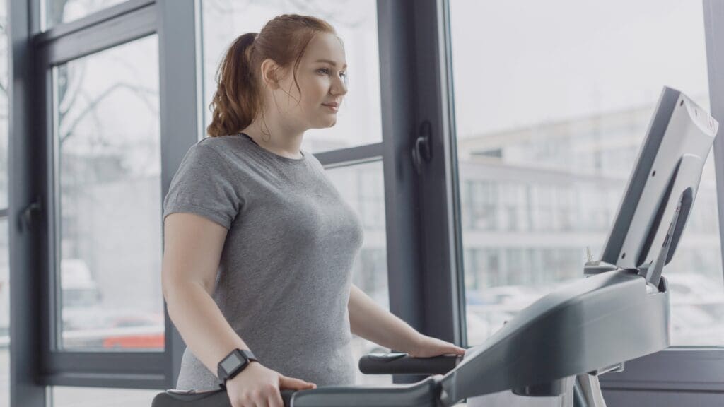 Person standing on a treadmill in front of a large window.