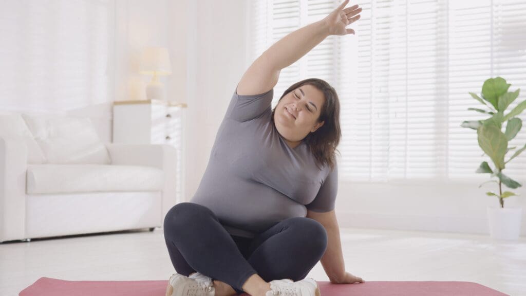 Person stretching their arm over their head while sitting on a yoga mat on the floor.